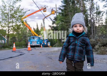 Porträt eines dreijährigen Jungen, der auf der Dorfstraße steht, als unscharfe Auftragnehmer bei der Arbeit im Hintergrund gesehen werden, die den elektrischen Sturm wiederherstellen. Stockfoto