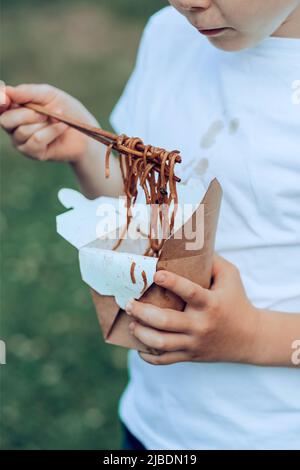 Das Kind, das unterwegs Sojasauce isst, gebratene Nudeln Essstäbchen. Schmutzige Sojasauce-Flecken auf einem weißen T-Shirt. Essen zum Mitnehmen. Im Freien Stockfoto