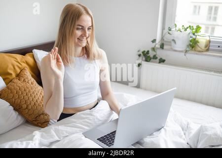 Bereit zum Lernen. Europäische Studentin sitzt auf dem Bett, bevor pc-Bildschirm machen Videoclip zu Tutor winken Hand Hallo. Stockfoto