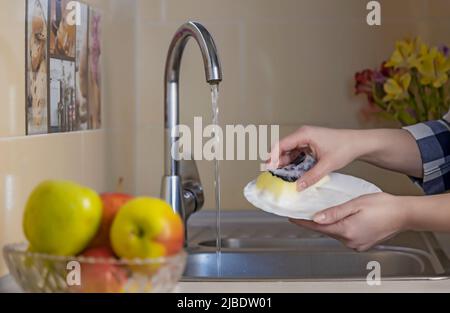 Frauenhänden waschen einen weißen Teller mit einem Schwamm unter fließendem Wasser im Spülbecken. Geschirr zu Hause waschen. Stockfoto