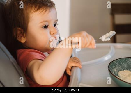 Versuchen Sie, den Haferbrei mit einem Löffel aus einer Schüssel zu essen. Kleiner Junge, der lernt, sich selbst zu ernähren. Lächelndes, glückliches Kind. Stockfoto