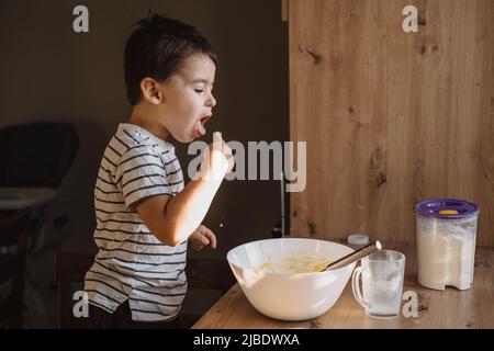 Junge saugt seinen Finger, um Schlagsahne zu probieren. Junge hilft in der Küche. Stockfoto