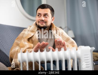 Mann mit einer Decke auf den Schultern und einer Tasse in der Nähe des Elektroheizgeräts Stockfoto