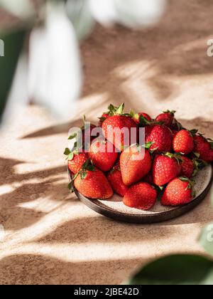 Erdbeeren in Teller auf braunem texturierten Hintergrund mit schönen Schatten, Blätter in der Unschärfe im Vordergrund Stockfoto