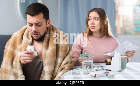 Kranker Mann, der neben seiner Frau am Tisch saß und ihm mit Medikamenten half Stockfoto