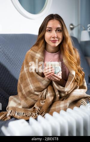 Frau mit einer Decke auf den Schultern und einer Tasse in der Nähe des elektrischen Heizgeräts Stockfoto