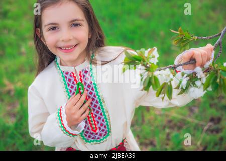 Glückliches schönes Mädchen genießen blühende Frühlingsbäume in Apfelgarten. Bulgarische Frau in Folklore-Kleid auf dem Feld. Stockfoto
