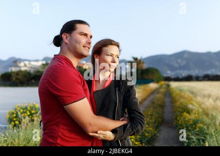 Porträt eines hübschen Paares mittleren Alters, das auf dem Copyspace zur Seite schaut und sich umarmt, während es im Sommerfeld auf Gras steht. Stockfoto