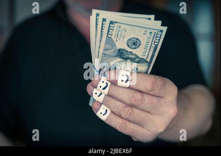 Ein erwachsener Mann reicht seine Hand mit $100 Rechnungen an die Kamera. Die ersten Fingerspitzen sind in ein weißes Band gewickelt. Lächelnde Gesichter sind Schmerz Stockfoto