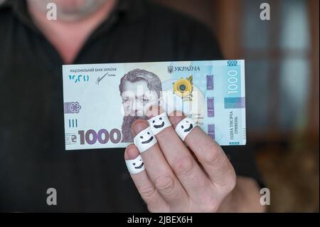 Ein erwachsener Mann hält Geld in der Hand. Eintausend ukrainische Griwnja. Die ersten Fingerspitzen sind in ein weißes Band gewickelt. Lächelnd Stockfoto