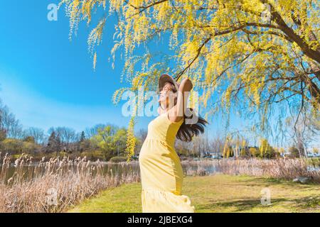 Glückliche asiatische Schwangere tanzen kostenlos im Natur Sommer Park trägt Sonnenhut und gelbes Kleid. Schwangerschaft Freude und sorglos Stockfoto