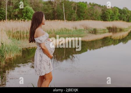 Natürliche Schwangerschaft Frau entspannen auf dem Land See tragen Umstandskleid für Naturspaziergang. Schwanger asiatische junge glücklich gesunde Mädchen im Freien Stockfoto