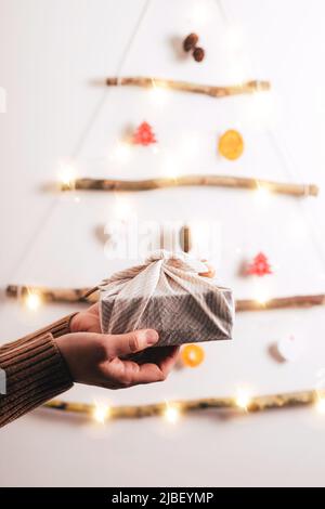 Frauenhände halten die furoshiki Geschenkbox gegen die Dekoration des Weihnachtsbaums aus trockenen Stäbchen, trockener Orange, Zapfen auf weißem Hintergrund. Null Stockfoto