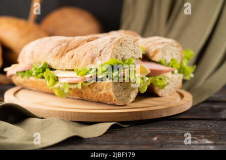 Fast-Food-Konzept, zwei frische U-Boot-Sandwiches mit Schinken, Käse, Speck, Salat, Gurken und Zwiebeln auf Holzschneidebrett Stockfoto