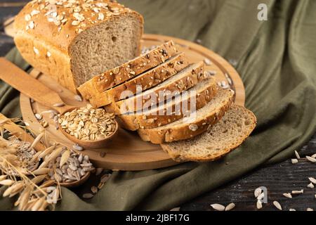 Mehrkornbrot auf einem Holzbrett mit Ohren aus Roggen und Hafer auf einem rustikalen Tuch im Hintergrund, hausgemachtes Backen Stockfoto