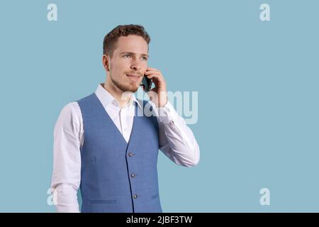 Serious jungen bärtigen Geschäftsmann in klassischen Anzug, der auf blauem Hintergrund Studios Porträt positioniert ist. Karriereerfolg Reichtum Geschäftskonzept. Auf den Kopierort ausfließen. Mobiltelefon wird gehalten Stockfoto