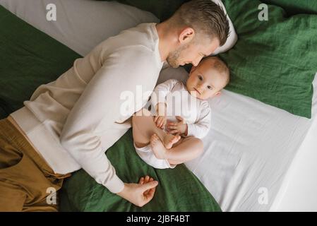 Ein hübscher, junger Vater liegt im Bett im Schlafzimmer und umarmt seinen liebend lachenden sechs Monate alten Jungen. Das Konzept der bedingungslosen Liebe und Vaterschaft Stockfoto