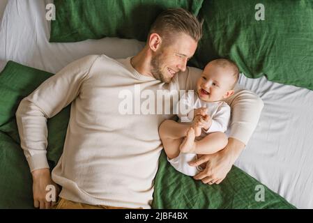 Ein hübscher, junger Vater liegt im Bett im Schlafzimmer und umarmt seinen liebend lachenden sechs Monate alten Jungen. Das Konzept der bedingungslosen Liebe und Vaterschaft Stockfoto