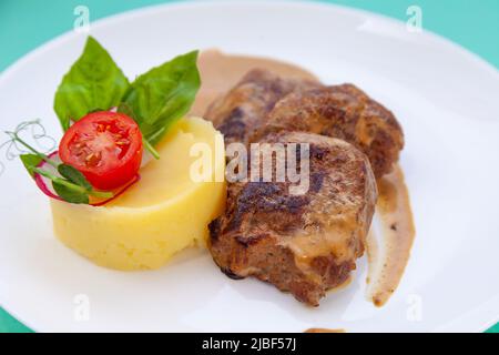 Fleischbällchen mit Kartoffelpüree auf einem weißen Teller. Fleisch in saurer Rahmsoße. Tomaten und Basilikum. Essen im Restaurant. Mittagspause. Medaillons. Stockfoto