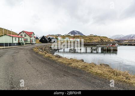 Eine Straße in Hofsós, Skagafjörður, Island, führt an blassgrünen Häusern mit roten Dächern vorbei auf dem Weg zum Hafen. Das Dorf liegt unter einem kegelförmigen Berg im Hintergrund. Ein hölzerner Steg ragt in die Bucht hinaus. Hofsós ist einer der ältesten Handelsposten des Landes, und heute leben dort etwa 170 Menschen. Zwischen 1850 und 1914 wanderten von hier aus etwa 20.000 Isländer nach Nordamerika aus. Stockfoto