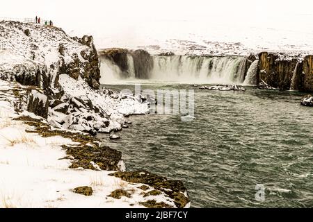 Goðafoss stürzt mit seiner vollen Breite über die Basaltklippen nahe Fosshóll, Bárðardalur, Nordisland, während winzige Besucher auf dem Aussichtsfelsen oben links im Bild stehen. Goðafoss, der "Wasserfall der Götter", ist etwa 30 Meter breit und 9 bis 17 Meter hoch, einer der wasserreichsten Wasserfälle des Landes und seit 2020 ein geschütztes Naturdenkmal. Nach isländischer Legende warf der Gesetzessprecher Þorgeir Ljósvetningagoði im Jahr 1000 seine Statuen der heidnischen Götter in den Wasserfall, nachdem das Land zum Christentum übergegangen war, was dem Wasserfall seinen Namen gab. Stockfoto