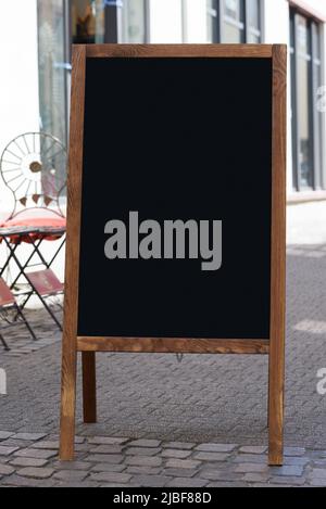 Leere hölzerne Sandwich-Tafel mit Tafel, auf der schmalen gepflasterten Straße mit Tisch und Stuhl im Hintergrund Stockfoto