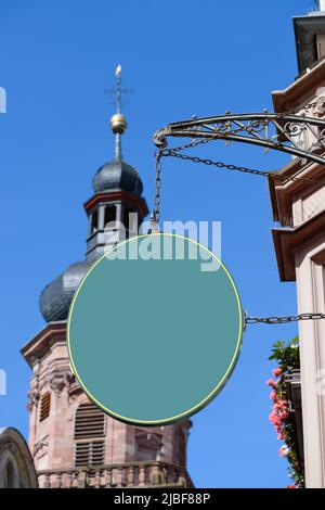 Blank grünes ovales Schild, das an einer alten Kette und einem geschmiedeten Vintage-Bogen an der Wand hängt, auf einer europäischen Straße mit Kirche im Hintergrund. Leere Plasti Stockfoto