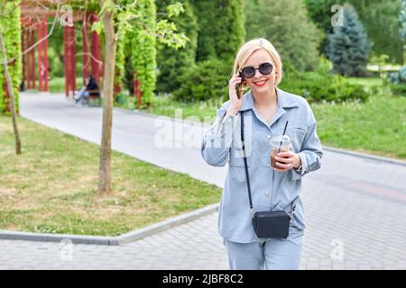 Stilvolle junge Blondine geht durch die Stadt und trinkt ein kaltes Getränk und telefoniert. Zeit nach der Arbeit Stockfoto