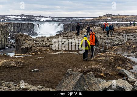 Mehrere Wanderer nähern sich über einen felsigen Pfad dem Aussichtspunkt über dem Wasserfall Selfoss an der Jökulsá á Fjöllum im Vatnajökull-Nationalpark im Nordosten Islands. Selfoss ist etwa 11 Meter hoch und liegt kaum einen Kilometer flussaufwärts von Dettifoss, erreichbar über einen markierten Weg entlang des Canyonrandes. Stockfoto