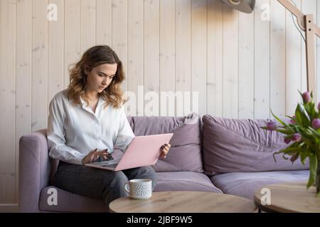 Seriöse junge Frau Geschäftsmann in weißem Hemd arbeitet zu Hause, auf gemütlichem Sofa und sorgfältig Typen Text, schaut durch Nachrichten. Online zu Hause arbeiten Stockfoto