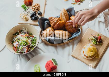 Frühstück im Hotel im Bett auf einem weißen Bettlaken, als eine Frau einen Brotkorb ergreift Stockfoto