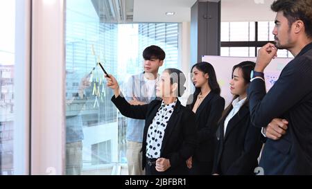 Fokussierte leitende weibliche Teamleiterin und diverse Geschäftsleute, die in einem modernen Büro zusammen mit Haftnotizen auf Glasfenstern Brainstorming durchführen Stockfoto