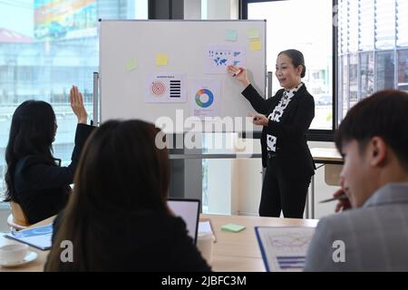 Selbstbewusste, reife Teamleiterin erklärt den Geschäftsplan und stellt jungen Mitarbeitern das Projekt auf einem Flipchart vor Stockfoto