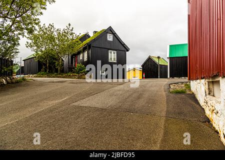 Schwarz geteerte Holzhäuser mit dicken Grasdächern säumen eine Straße in der Altstadt von Tórshavn, Färöer-Inseln. Rechts schließt sich ein rotes Lagerhaus mit einem grünen Wellblechdach an. Stockfoto