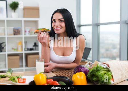 Junge Frau, die in der Küche steht und sich an der Holztheke lehnt, hat ein Sandwich und ein Glas Milch zum Mittagessen Stockfoto