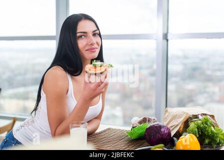 Nahaufnahme einer Frau, die in der Küche steht, sich auf einen Holztisch lehnt und einen Snack iß. Lächelndes Mädchen pflegt einen gesunden Lebensstil Stockfoto