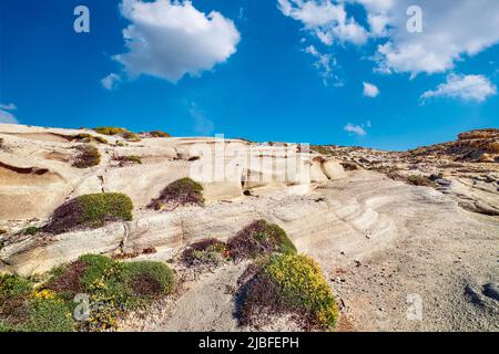 Sommerlandschaft von Felsen am Sarakiniko Strand, Ägäis, Insel Milos, Griechenland. Keine Menschen, leere Klippen, Sonnenschein, blauer Himmel und Wolken, etwas Gras Stockfoto