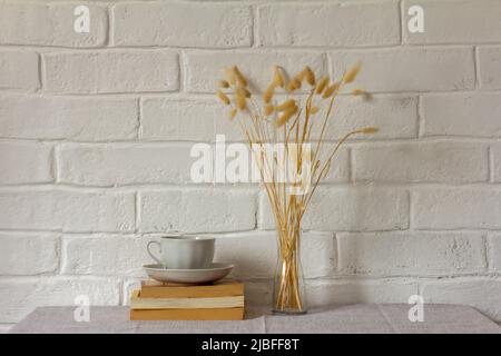 Eine Tasse Kaffee, Bücher und eine Glasvase mit getrockneten Lagurusblüten. Beigefarbene Tischdecke aus Leinen. Dorfhaus Stockfoto