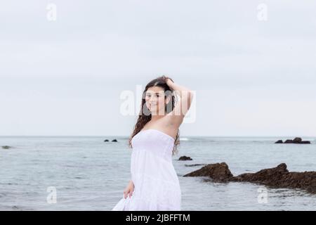 Junge Brünette Frau in weißem Kleid posiert am Meer Stockfoto