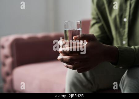 Hände eines jungen, beunruhigten afroamerikanischen Mannes, der ein Glas Wasser in der Hand hält, während er im Büro seines Psychoanalytikers oder Psychologen auf der Couch sitzt Stockfoto