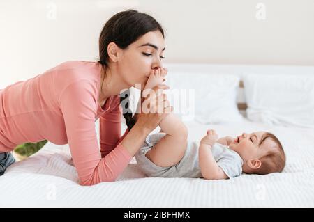 Fürsorge der Mutter. Liebevolle Mutter küsst kleine Füße ihres Säuglings und genießt es, sich zu Hause mit dem Kleinkind zu verbinden Stockfoto