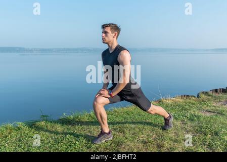 Ein junger, frecher Mann streckt die Beine im Freien und macht einen Ausfallschritt nach vorne Stockfoto