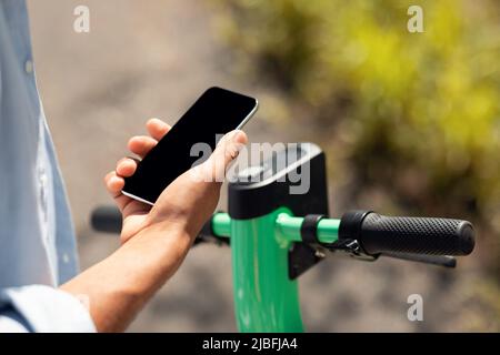 Konzept der Elektromobilität. Schwarzer Mann entriegelt E-Scooter mit seinem Smartphone mit schwarzem leerem Bildschirm, Modell für Design Stockfoto