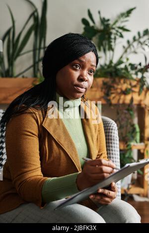 Junge, seriöse weibliche Counselin hört sich die Patientenerzählung an und macht sich Notizen in Dokumenten, während sie während der Sitzung im Sessel sitzt Stockfoto