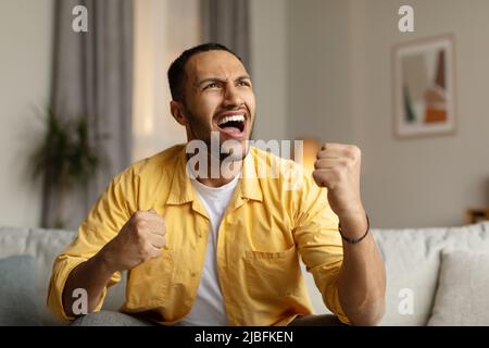 Emotionaler junger schwarzer Mann, der schrie, ja vor Freude gestikulte, den Sieg feierte, zu Hause auf dem Sofa saß Stockfoto