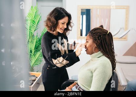 Fokussierte weibliche Visagiste, die während der Make-up-Session im Studio mit Fingern die Grundlage auf die Wange einer schwarzen Frau mit geflochtenen Haaren verschmiert Stockfoto