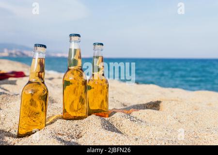 An sonnigen Tagen im Resort werden Glasflaschen mit kaltem Bier auf Sand auf unscharfem Hintergrund des Meeres und des blauen Himmels gelegt Stockfoto