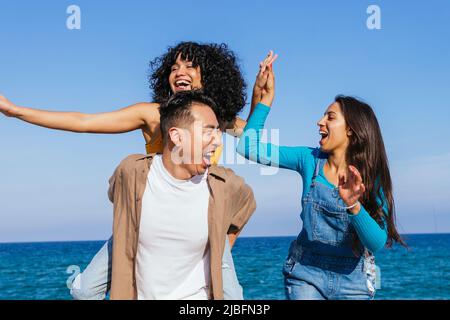 Fröhliche hispanische Frau in legerer Kleidung lachend und geben hohe fünf zu schwarzen Freundin sitzen auf der Rückseite des asiatischen Freund gegen Meer und blau sk Stockfoto