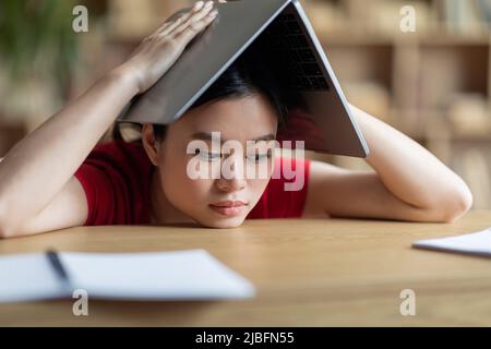 Müde frustriert teen asiatische Mädchen macht Dach mit Buch auf ihrem Kopf in der Bibliothek oder Raum Interieur Stockfoto