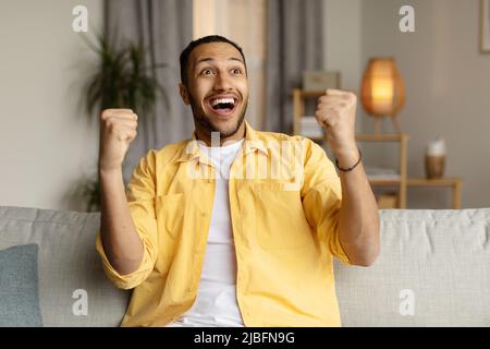 Überglücklich junger schwarzer Mann, der vor Aufregung schreit, ja gestikuliert, zu Hause auf der Couch sitzt Stockfoto